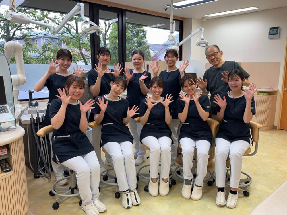 酒井歯科医院の診療室で、ネイビーのユニフォーム姿の歯科スタッフ9名と院長が笑顔で両手を広げて並ぶ集合写真